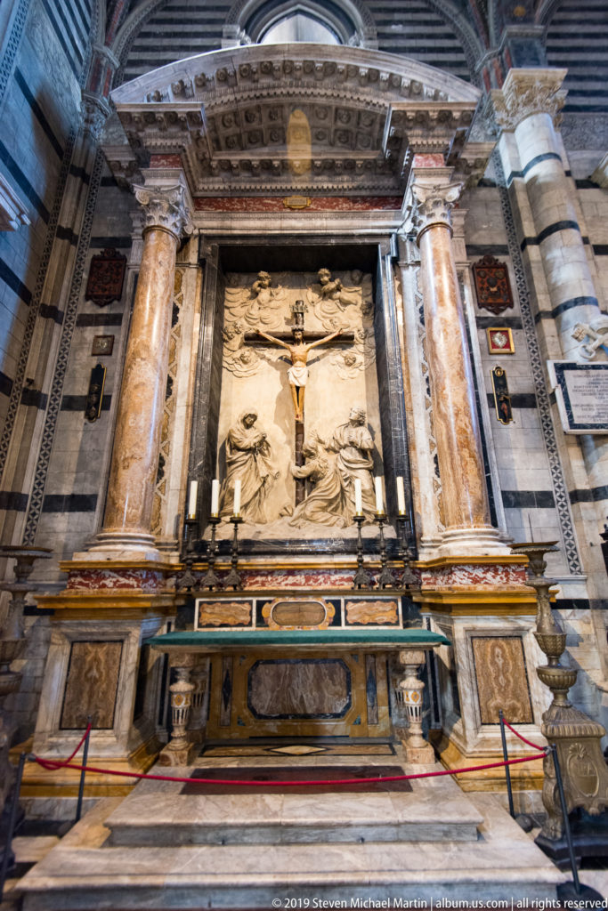 A crucifixion painting hangs above candles and a marble altar between two tall columns.