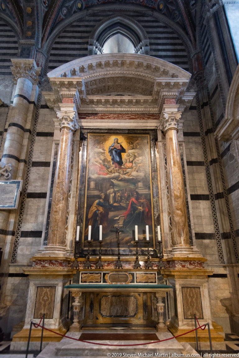 A framed religious painting rises above candles and an ornate marble altar between stone columns.