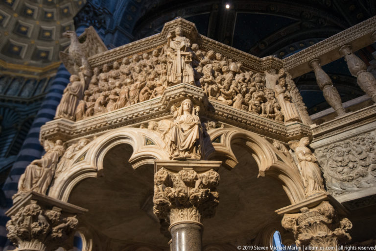 Pale marble figures project from the upper edge of the cathedral pulpit above carved arches.