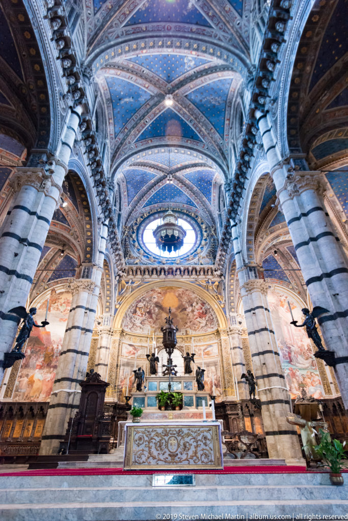 The altar and painted apse appear between striped piers beneath Siena Cathedral’s blue vault.