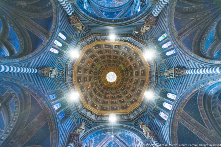 A gilded geometric ceiling bay converges on a small blue-white medallion above the cathedral nave.