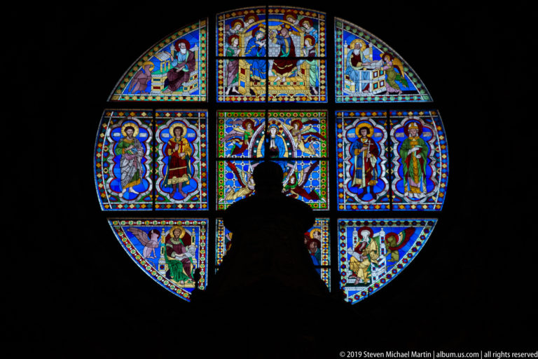 A visitor’s silhouette partly blocks a colorful round stained-glass window in a dark cathedral interior.