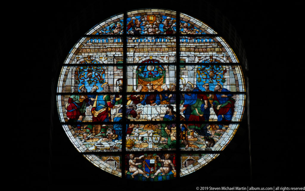 A circular stained-glass window filled with colorful narrative figures and architectural scenes.