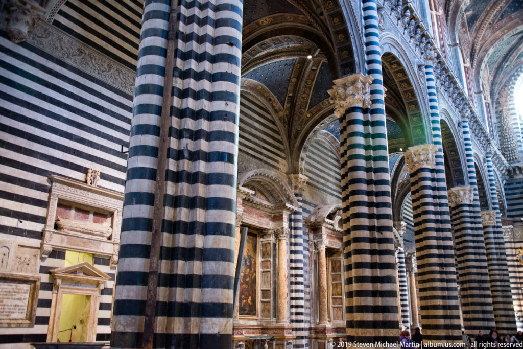 A side aisle recedes between striped columns toward a bright window and a framed religious image.