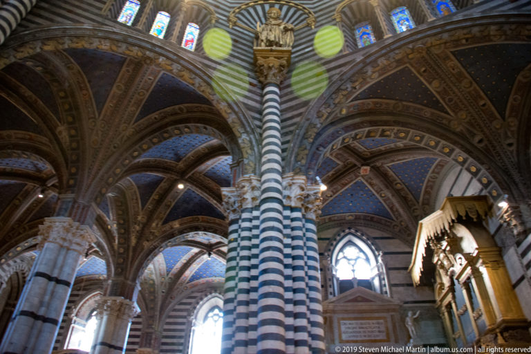 Striped piers and crossing arches rise toward blue and gold vaults inside the Duomo di Siena.