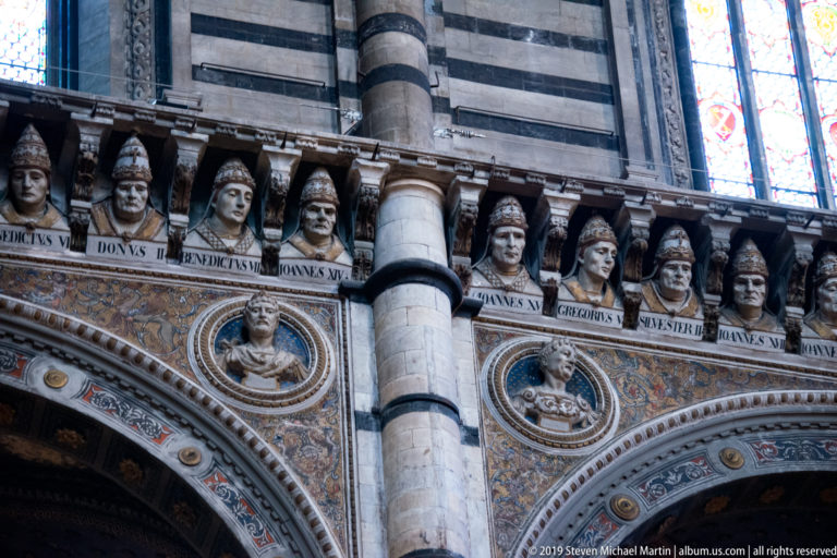 Carved papal busts and name inscriptions run above a decorated interior arch beside a striped pier.
