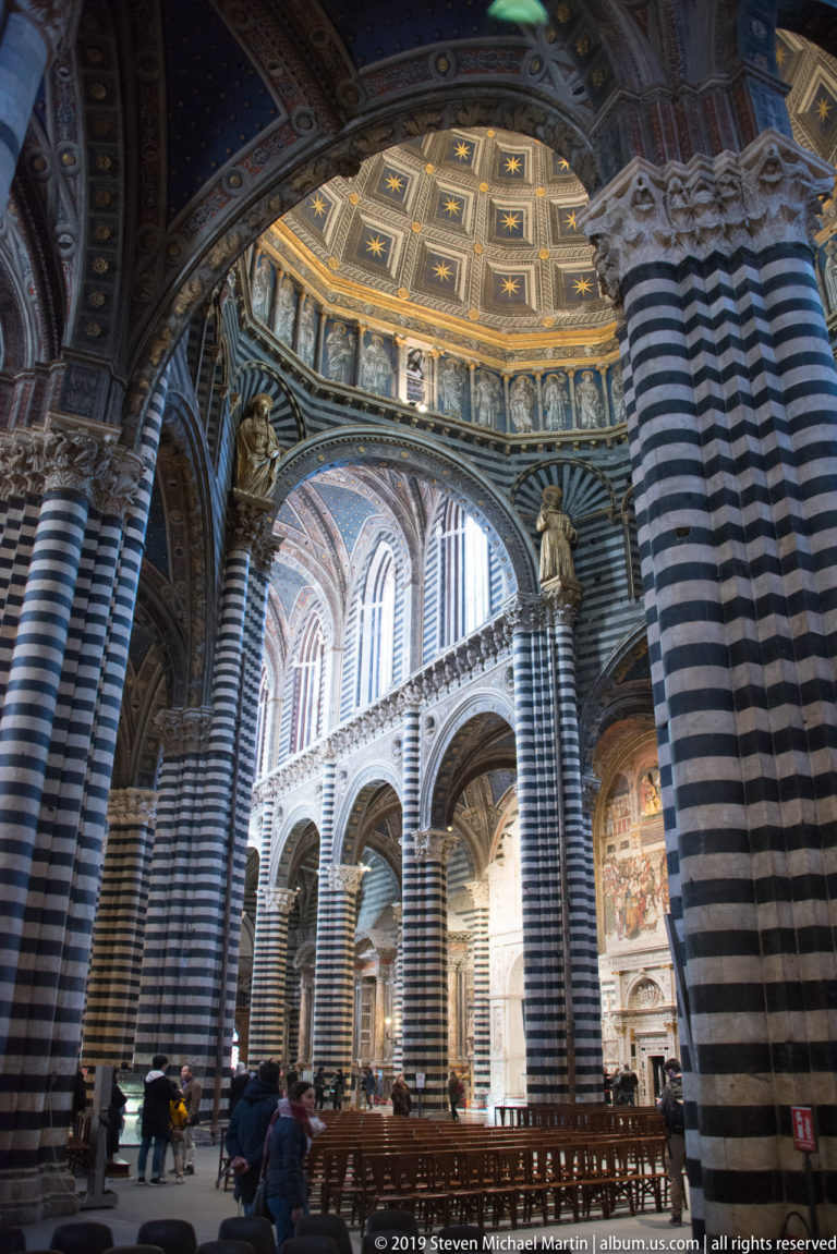 Visitors stand among benches beneath the striped piers and high arches of Siena Cathedral.