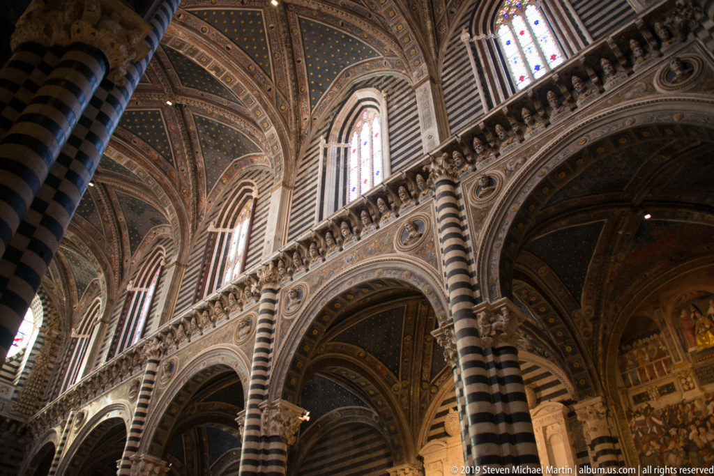 Upward view of striped stone piers and pointed arches beneath a blue, gold-starred cathedral vault.
