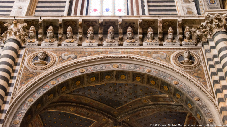 Stone busts of popes with name panels in a horizontal gallery on Siena Cathedral’s façade.