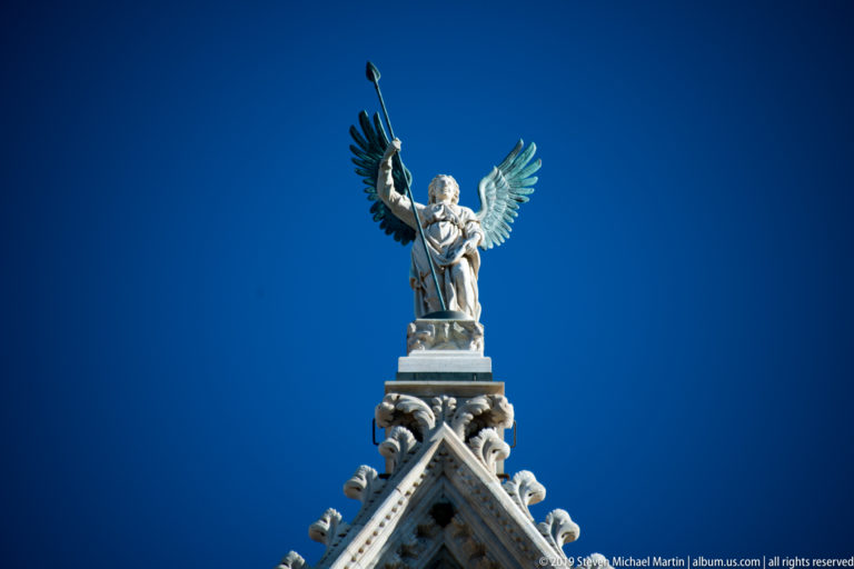 A winged stone figure holding a staff stands above a pointed cathedral gable.
