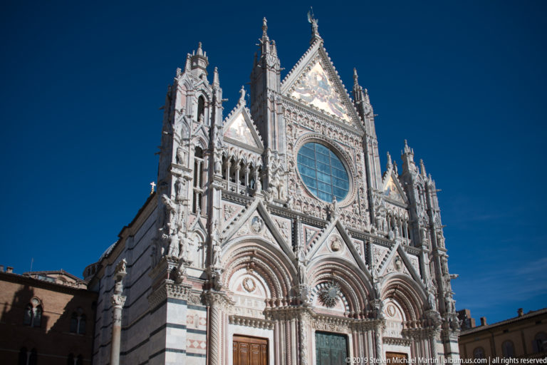 Upward view of Siena Cathedral’s striped marble west façade, with pointed gables, pinnacles, and a large central oval window.