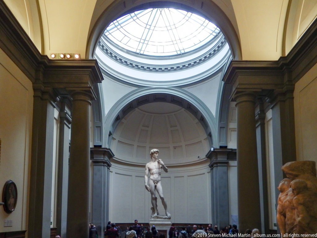 Long gallery view toward Michelangelo’s David beneath the glazed dome and Tribune, with visitors gathered below and columns framing the axis.