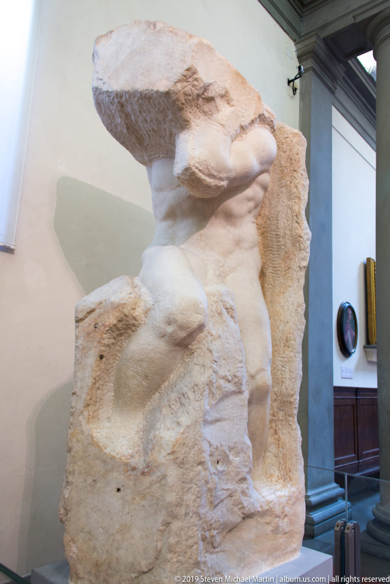 Contextual side view of Michelangelo’s Atlas Slave emerging from a tall rough marble block inside the museum gallery.