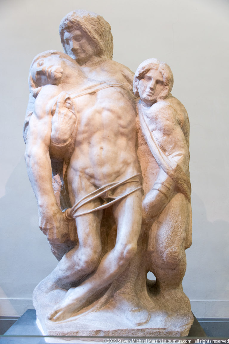 Broader view of a sculptural group with a collapsed male figure supported by attendants on a rough integral base.