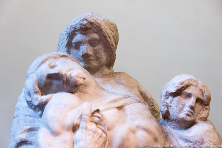 Close sculptural group with a reclining male figure supported by two attendants whose faces rise behind his shoulders.