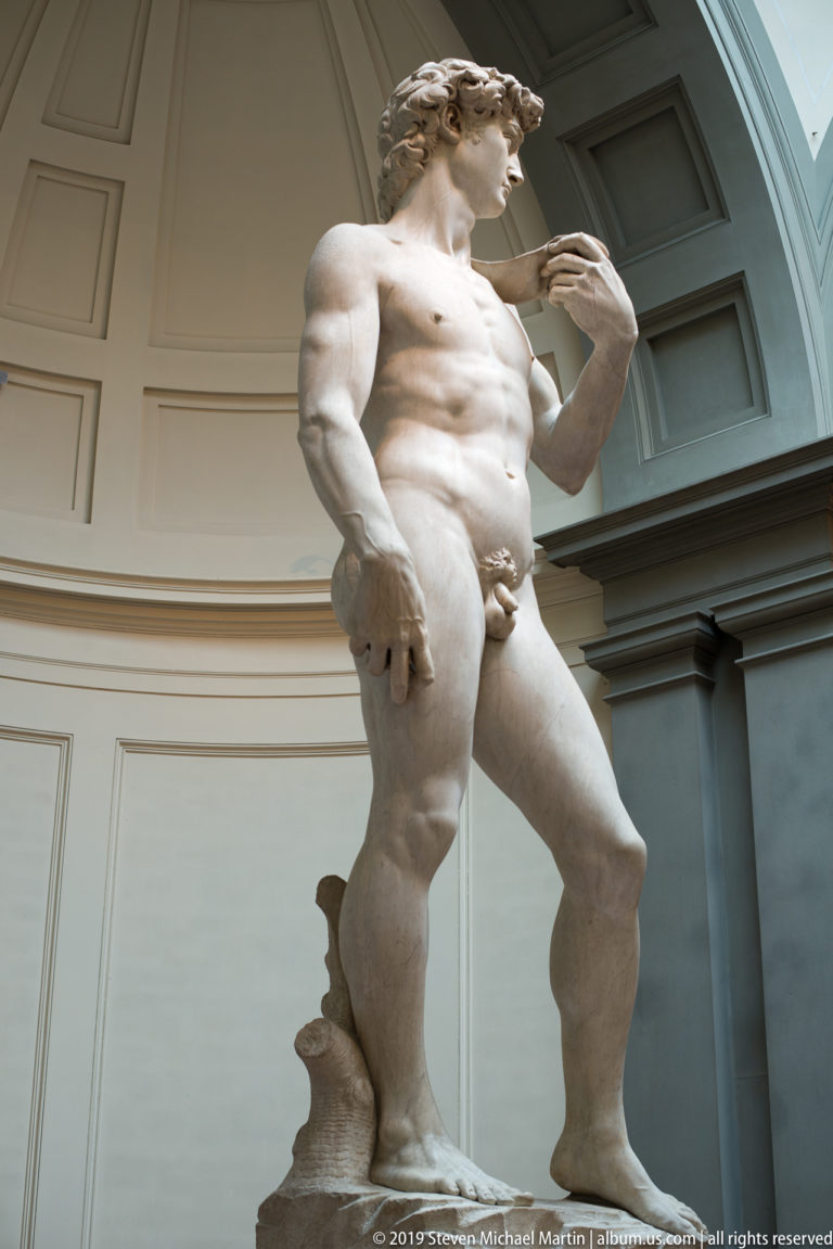 Right three-quarter low view of Michelangelo’s David with lowered hand prominent, head turned, and sling hand raised near the shoulder.