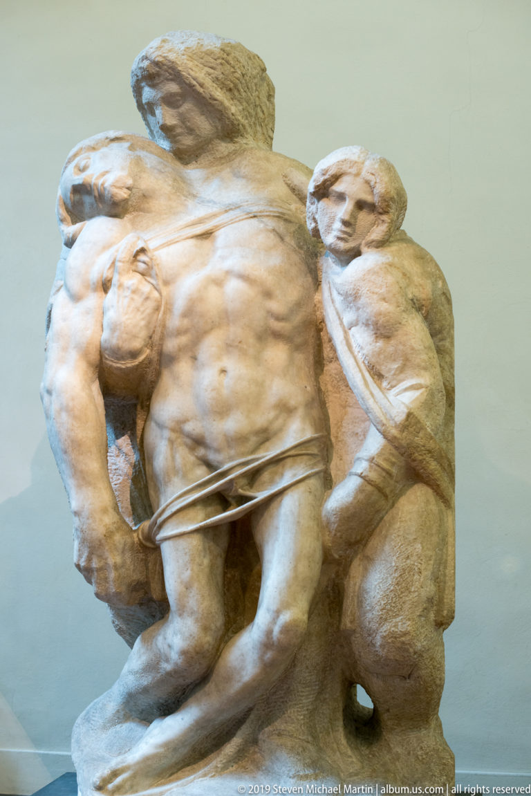Michelangelo’s Bearded Slave with muscular torso, bowed bearded head, and a less-finished figure or mass attached behind.