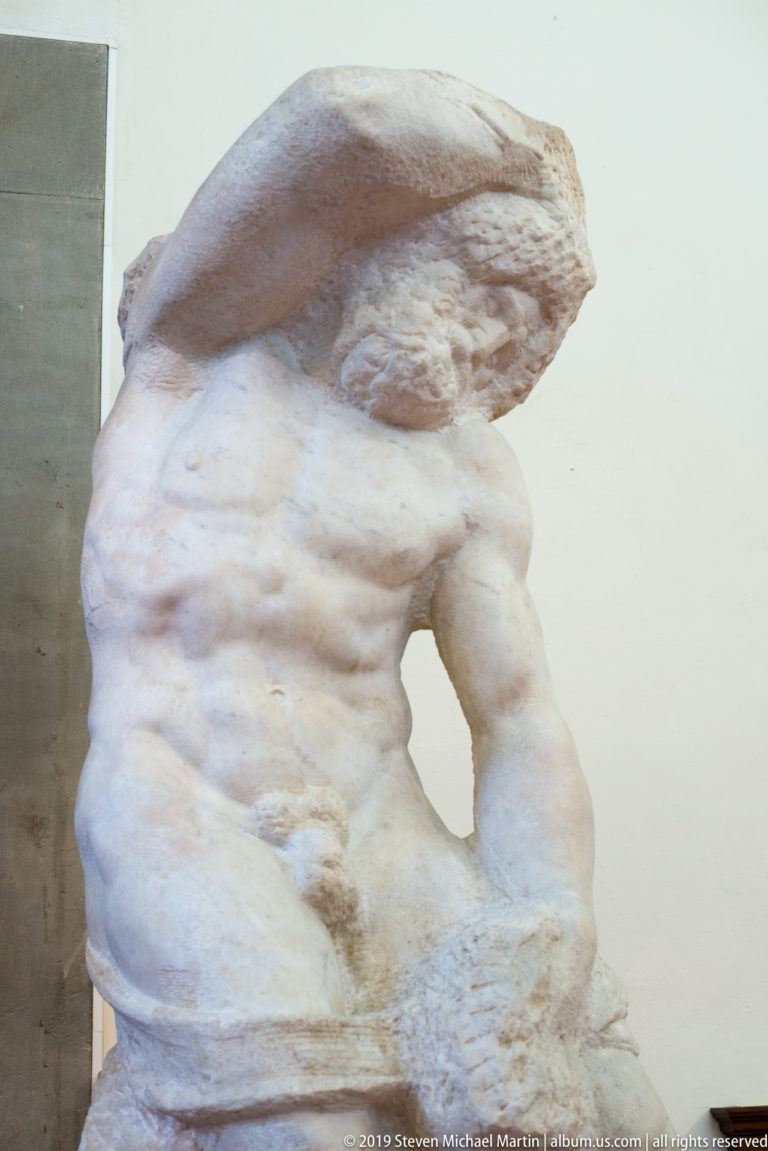Frontal view of an unfinished Michelangelo captive with modeled chest and abdomen emerging from rough stone around the head and lower body.
