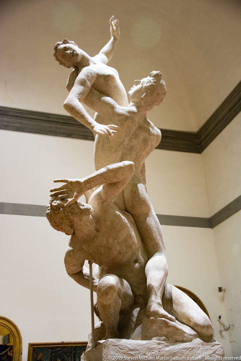 Side view of a dramatic sculpture group with a kneeling bearded figure below and two twisting nude figures rising above.