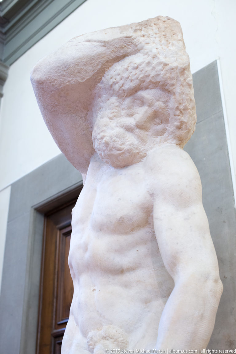 Low side view of Michelangelo’s Atlas Slave with torso and arm emerging below a large rectangular mass of rough marble.