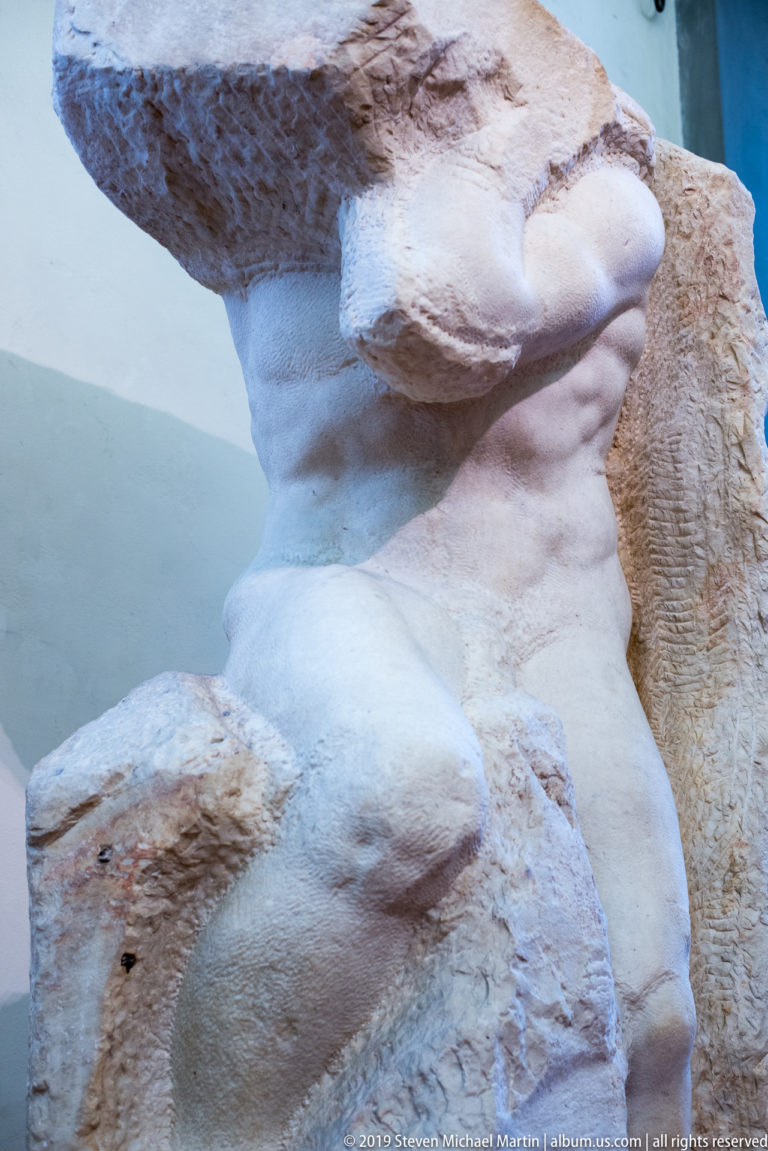 Close torso view of Michelangelo’s Atlas Slave, with modeled chest and abdomen emerging beneath a large rough marble mass.