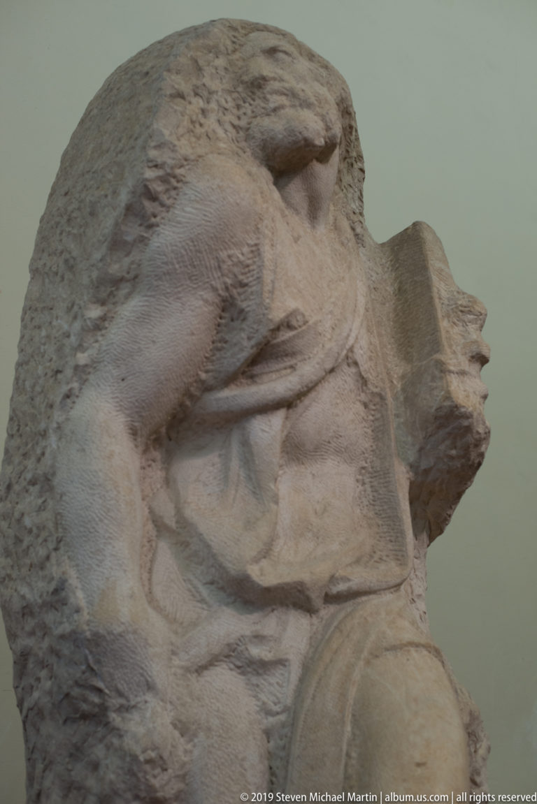 Side view of an unfinished Michelangelo captive figure partially emerging from a rough marble block, with bent torso and projecting profile.