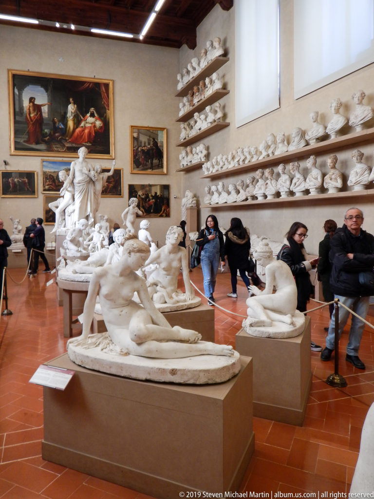 Wide museum interior filled with pale sculptures, reclining groups, portrait busts on shelves, visitors, and paintings on the walls.