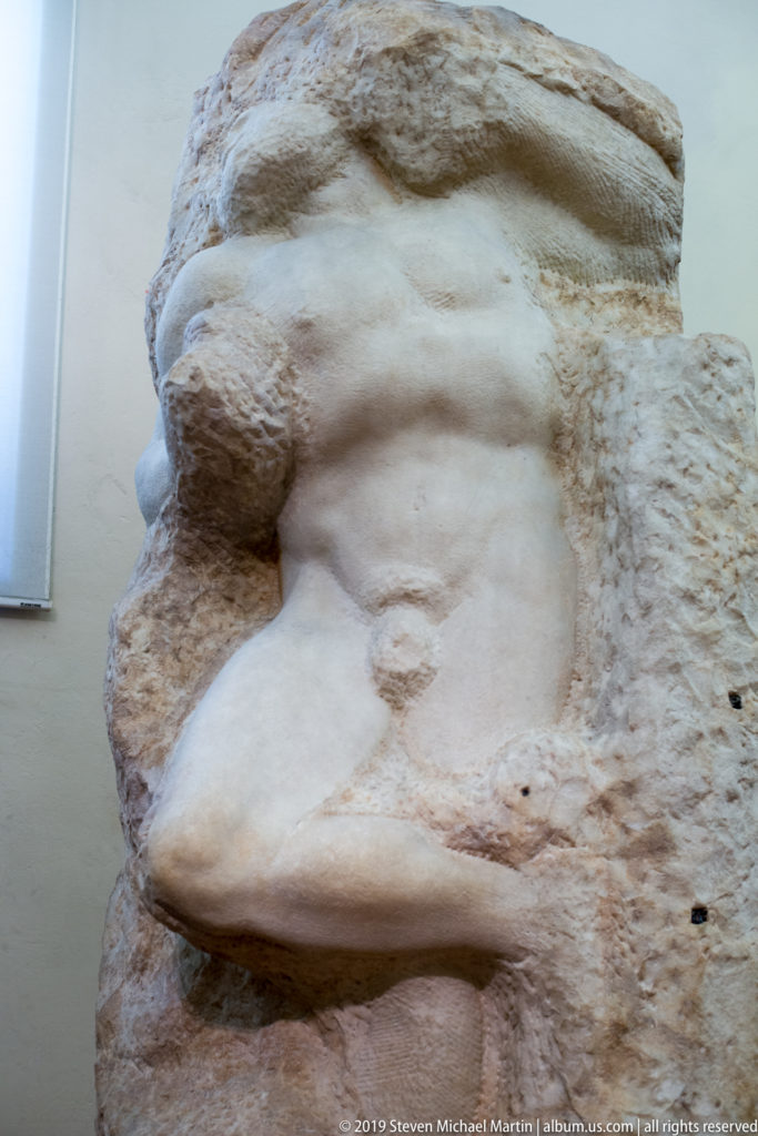 Close view of Michelangelo’s Atlas Slave emerging from a rough marble block beneath a massive uncarved section above the shoulders.