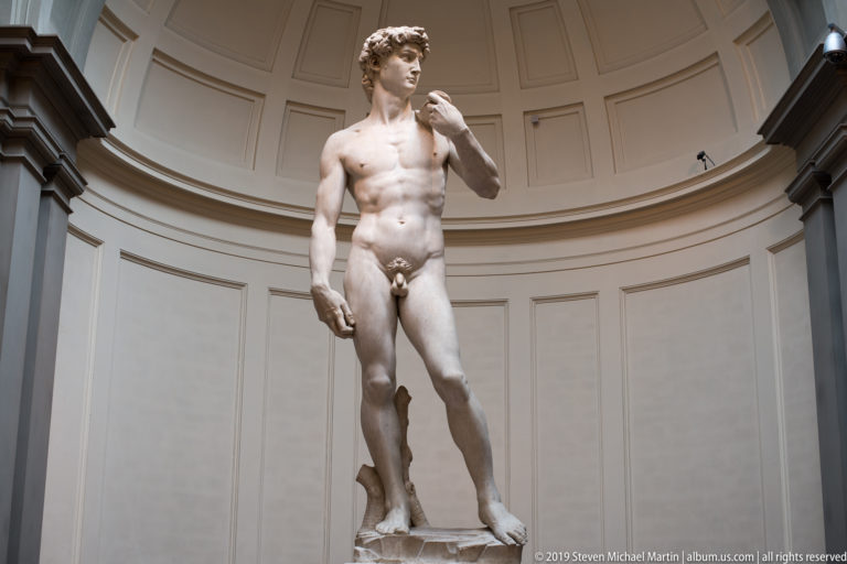 Centered full-length view of Michelangelo’s David beneath the curved Tribune wall, with head turned and left hand raised.