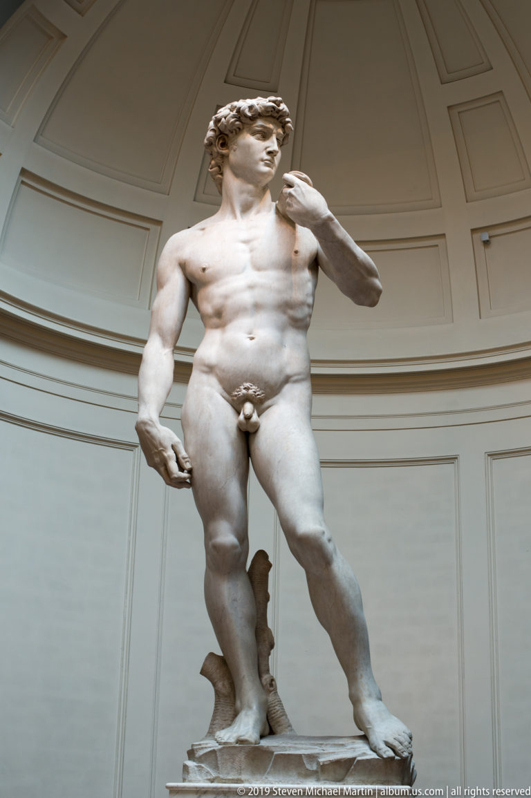Three-quarter low view of Michelangelo’s David with head turned, sling at the shoulder, and weight shifted onto one leg.