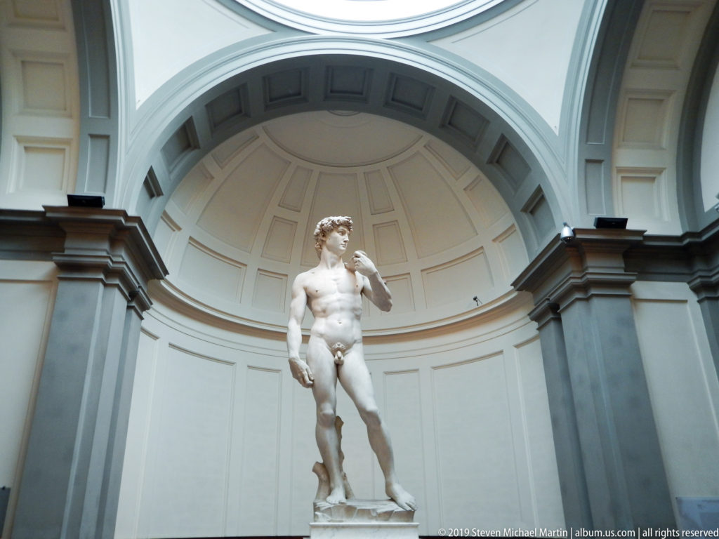 Wide symmetrical view of Michelangelo’s David centered beneath the coffered half-dome and monumental arches of the Tribune.