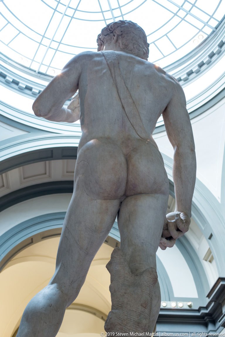 Rear view of an unfinished Michelangelo captive figure beneath the curved glazed roof, with rough stone crossing the back.