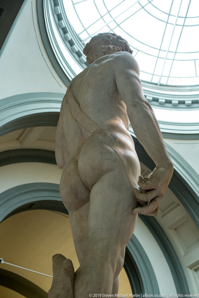 Low rear view of an unfinished Michelangelo captive figure beneath a glazed museum ceiling, showing massive back and rough stone surfaces.