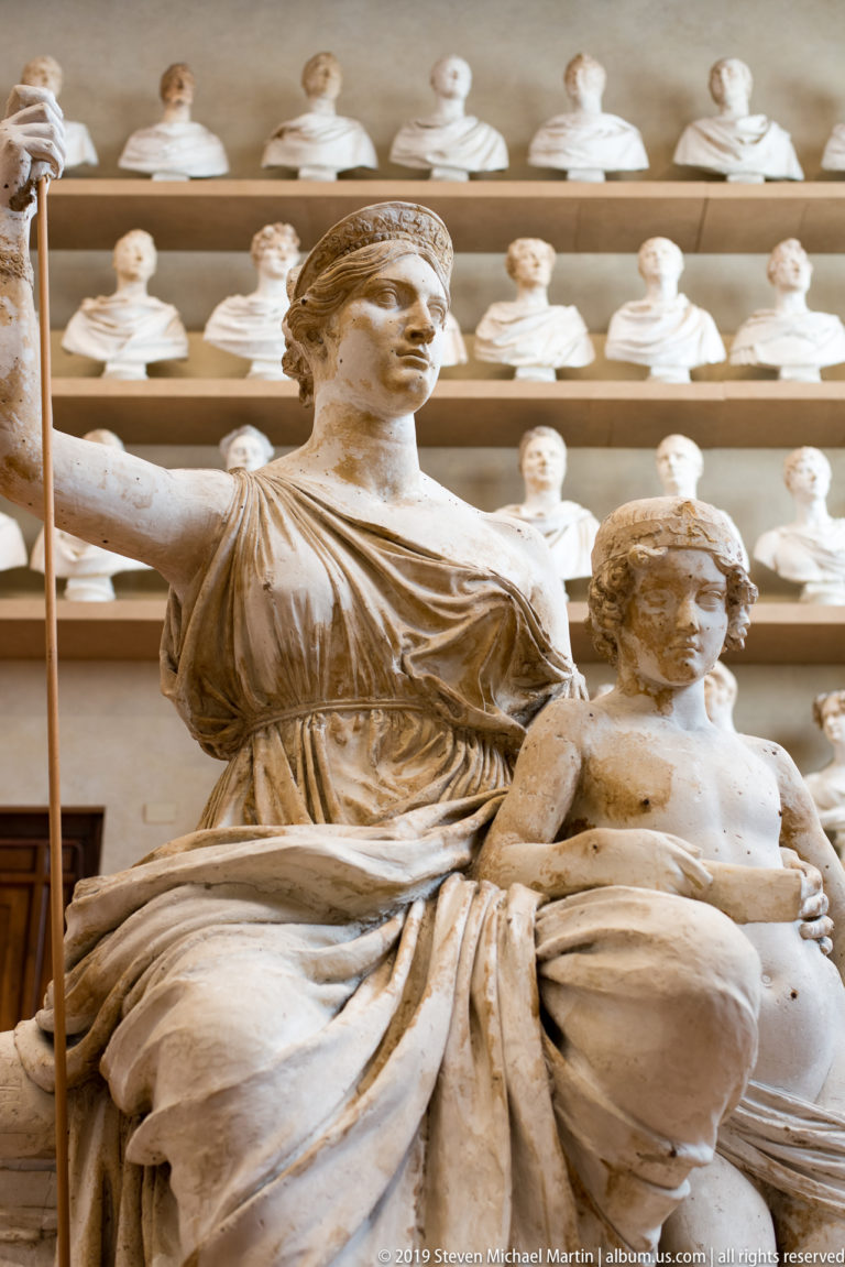 Seated draped female sculpture with one arm raised and a child leaning beside her, backed by rows of portrait busts.