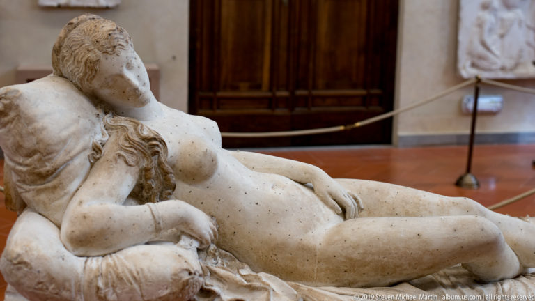 Reclining bearded male sculpture extending diagonally across the frame, with one hand near his head and gallery displays behind.