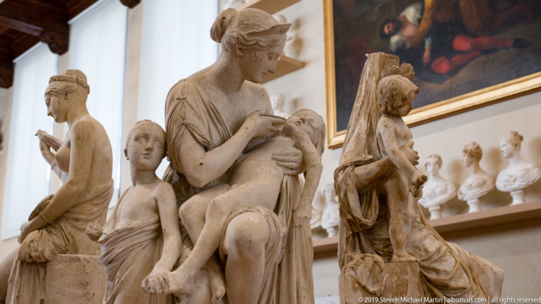 Cluster of pale figure sculptures in a study gallery, including a standing woman bending toward a child and seated figures nearby.