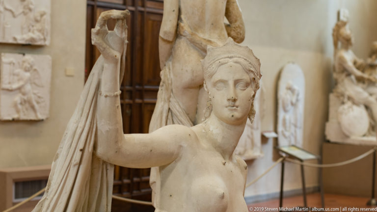 Waist-up female sculpture with one arm raised beside drapery, surrounded by other works in the museum gallery.