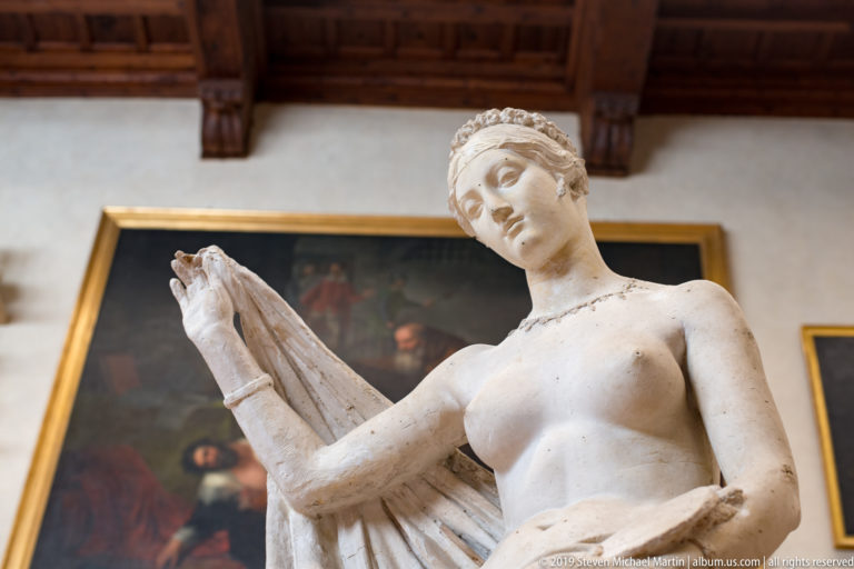 Low-angle view of a standing female sculpture lifting drapery, with a dark framed painting and wooden ceiling behind.