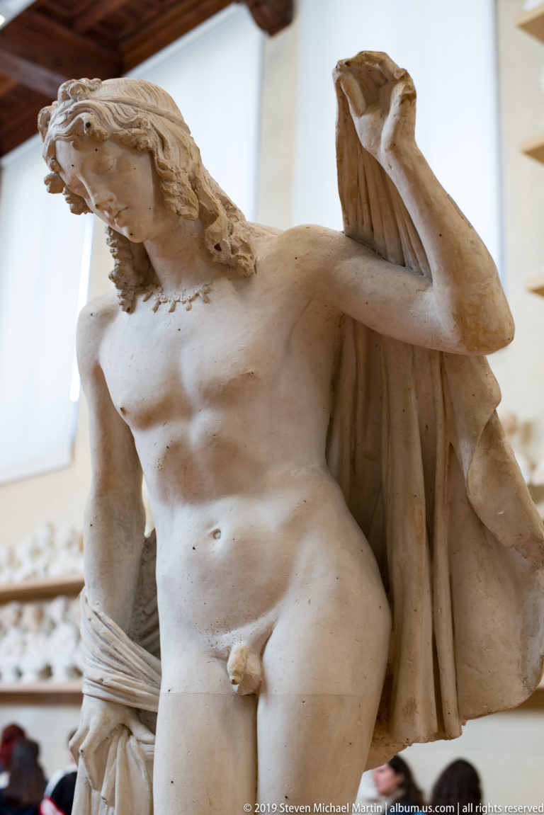 Fragmentary male nude sculpture with missing head, raised damaged arm, drapery behind the torso, and visible surface losses.