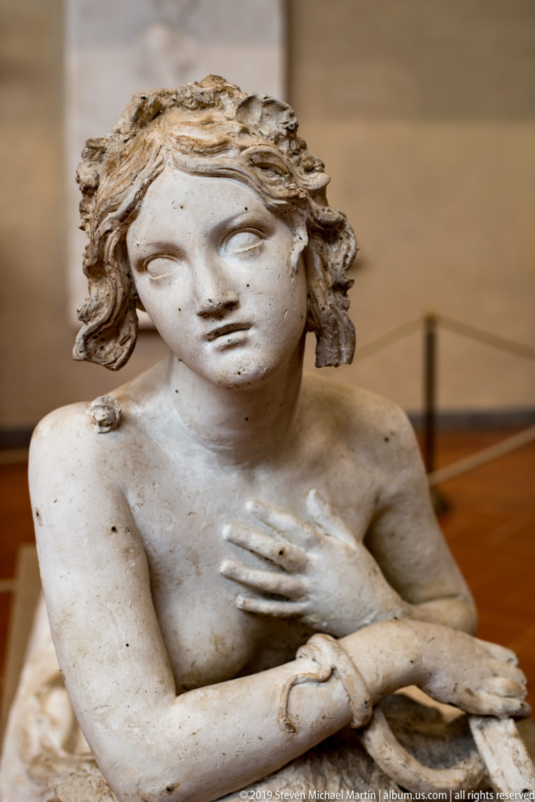 Close view of a reclining female sculpture with wavy hair, one hand on her chest and the other extended forward.