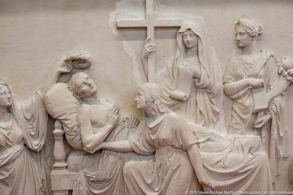 Stone or plaster relief showing a reclining figure surrounded by women beneath a large upright cross.