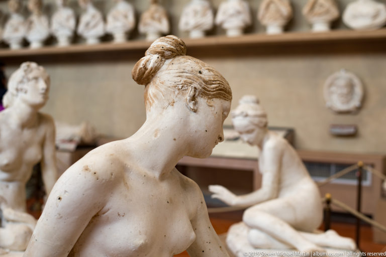 Profile of a pale female sculpture with hair in a high bun, with other seated and standing figures blurred behind.