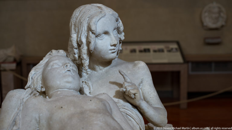 Close view of one female sculpture supporting another reclining figure whose face turns upward toward her.