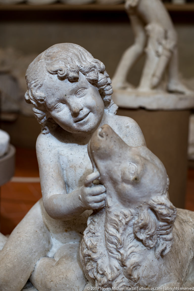 Child embraces the head of a reclining adult sculpture, with intertwined arms and textured hair shown in close view.