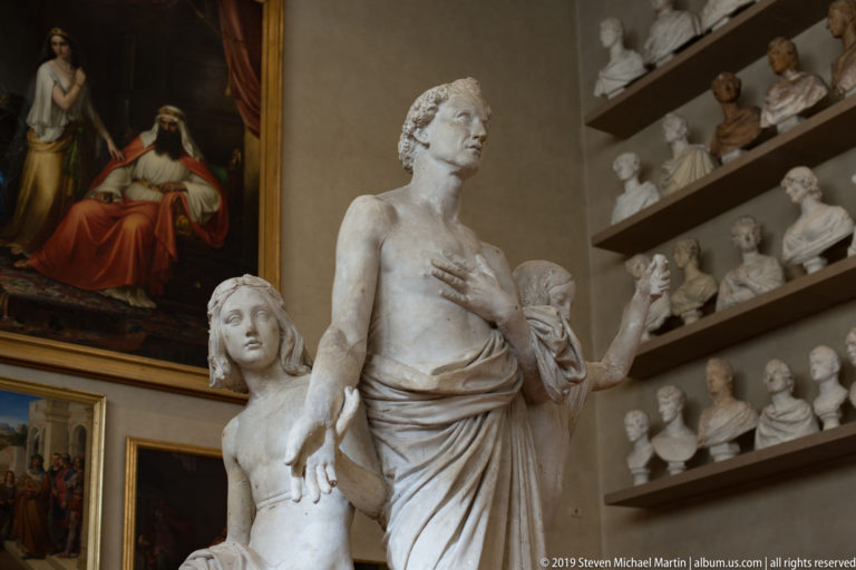 Large draped female sculpture with one hand at her chest and another figure beside her, backed by shelves of portrait busts.