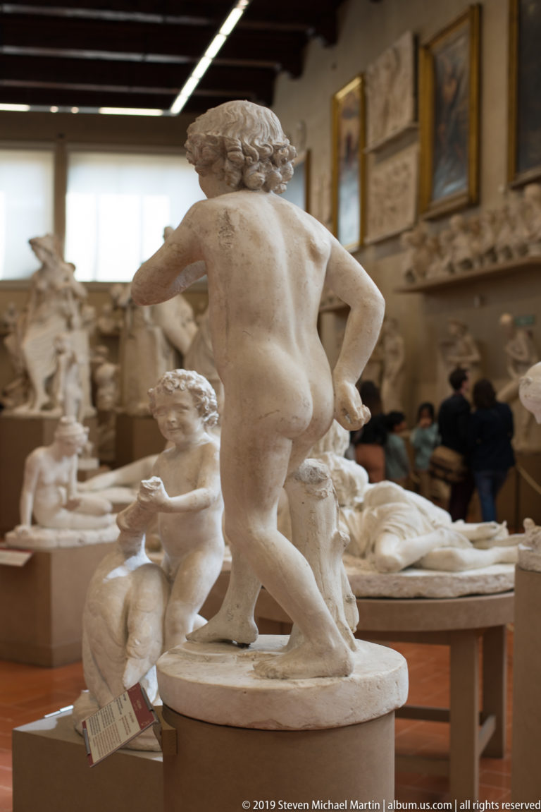 Rear view of a nude male sculpture on a round base, with a smaller child figure and crowded sculpture gallery behind.
