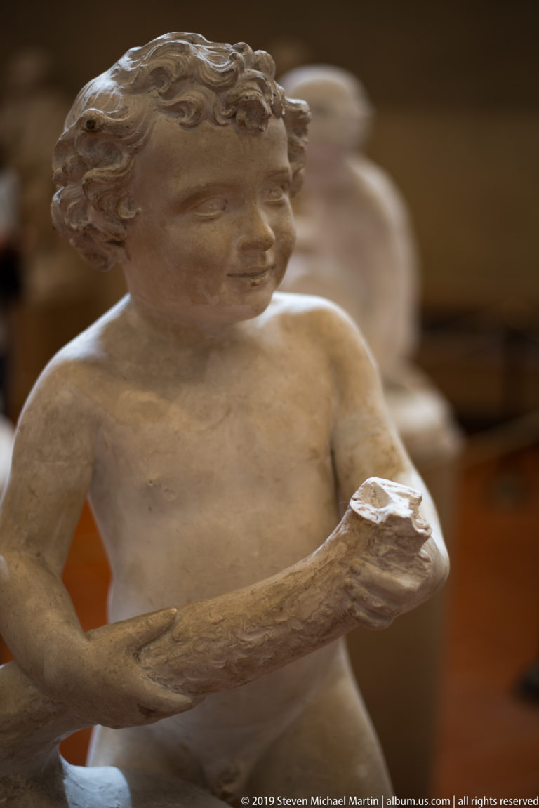 Close view of a nude male sculpture with curled hair, turned head, and both hands holding a rectangular object.