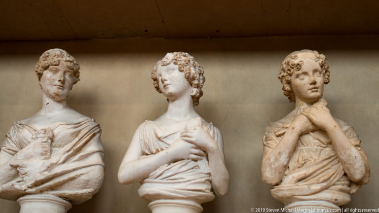 Three pale male portrait busts with curled hair and draped clothing displayed side by side on a museum shelf.