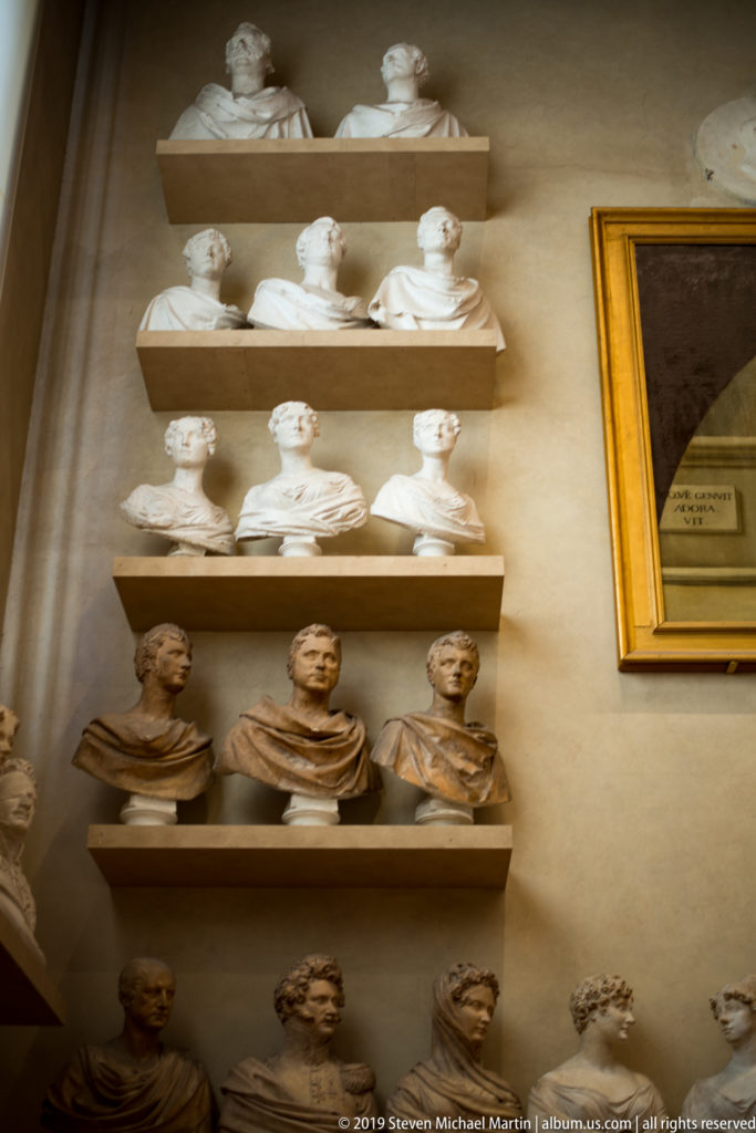 Oblique museum view of multiple rows of portrait busts arranged closely on wall-mounted shelves.