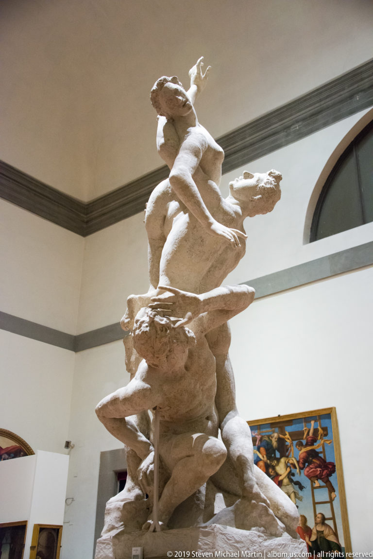Low-angle view of a multi-figure sculpture with intertwined nude bodies, raised arms, and a crouching figure at the base.